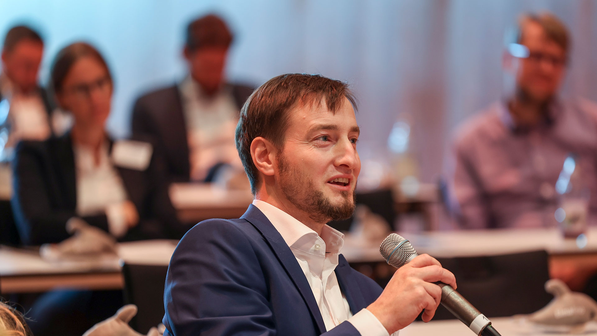 Frage aus dem Publikum. Mann mit Mikrofon sitzend, umrandet von Plenum.  